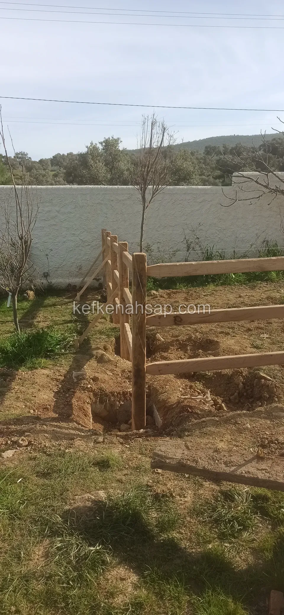 Three horizontal rails fixed to thick timber posts, with dug-up soil around and a white wall behind