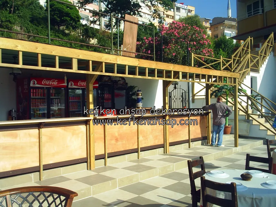 Satranç desenli teras zemininde, kirişli pergolanın altında uzanan ahşap bar tezgahı; yanda merdiven ve bir kişi