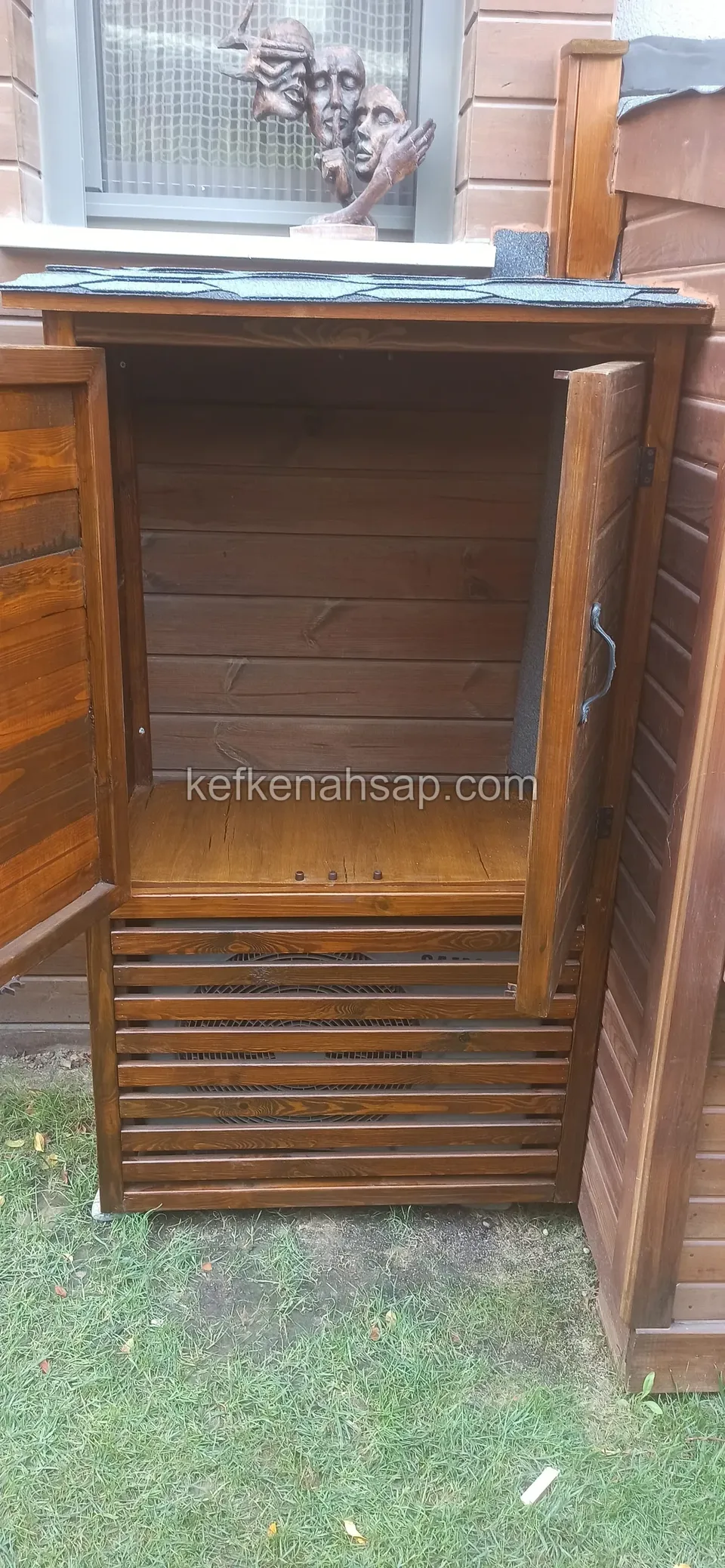 Pencere altındaki küçük ahşap dolabın kapakları açık, içinde yatay tahta kaplı iç yüzey ve alt raf, çimenlik üzerinde