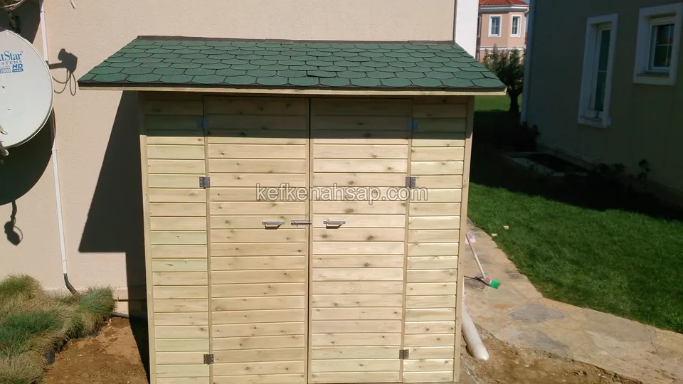 Yeşil shingle çatılı, yatay tahtalı açık renk ahşap depo; çift kapaklı, bina duvarının önünde duruyor