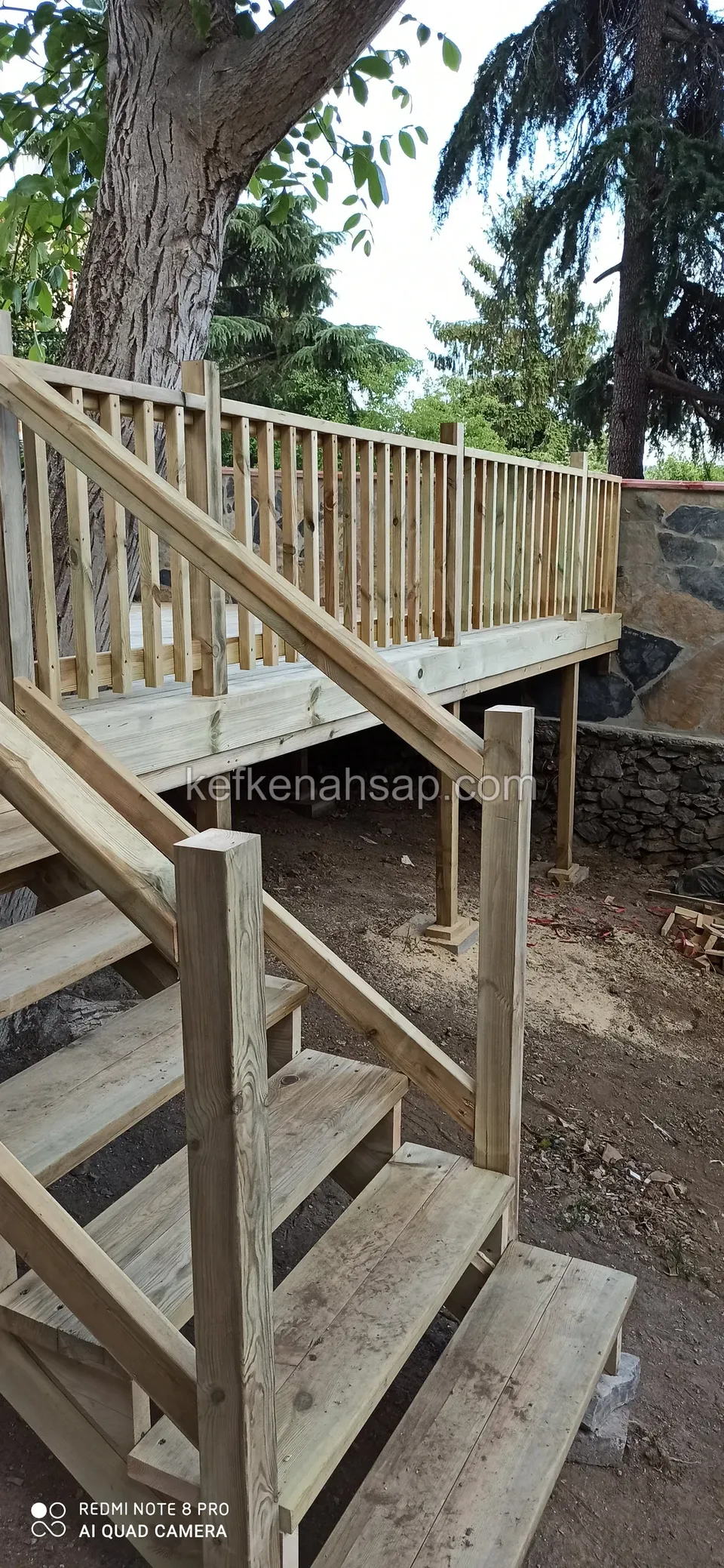 Ağacın altında yükseltilmiş ahşap deck, çıkışında korkuluklu ahşap merdiven; arkada taş duvar