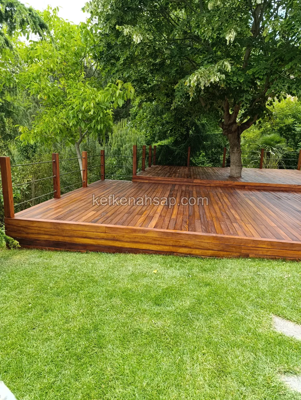 Two-level dark timber deck built around a large tree at the edge of a lawn
