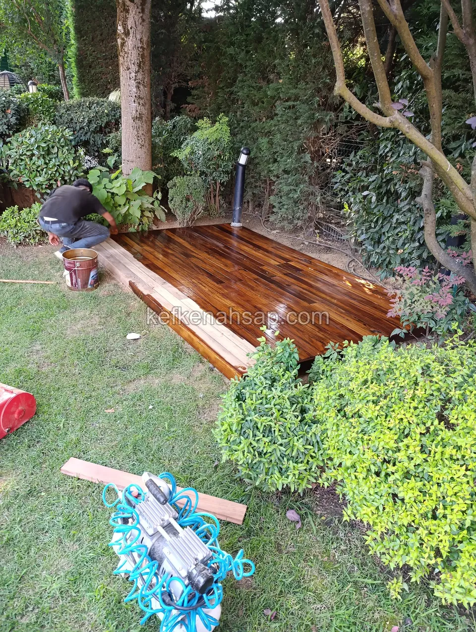 Newly laid, freshly finished dark deck beside a tree in a lawned garden, with a craftsman, a paint tin and tool bags nearby