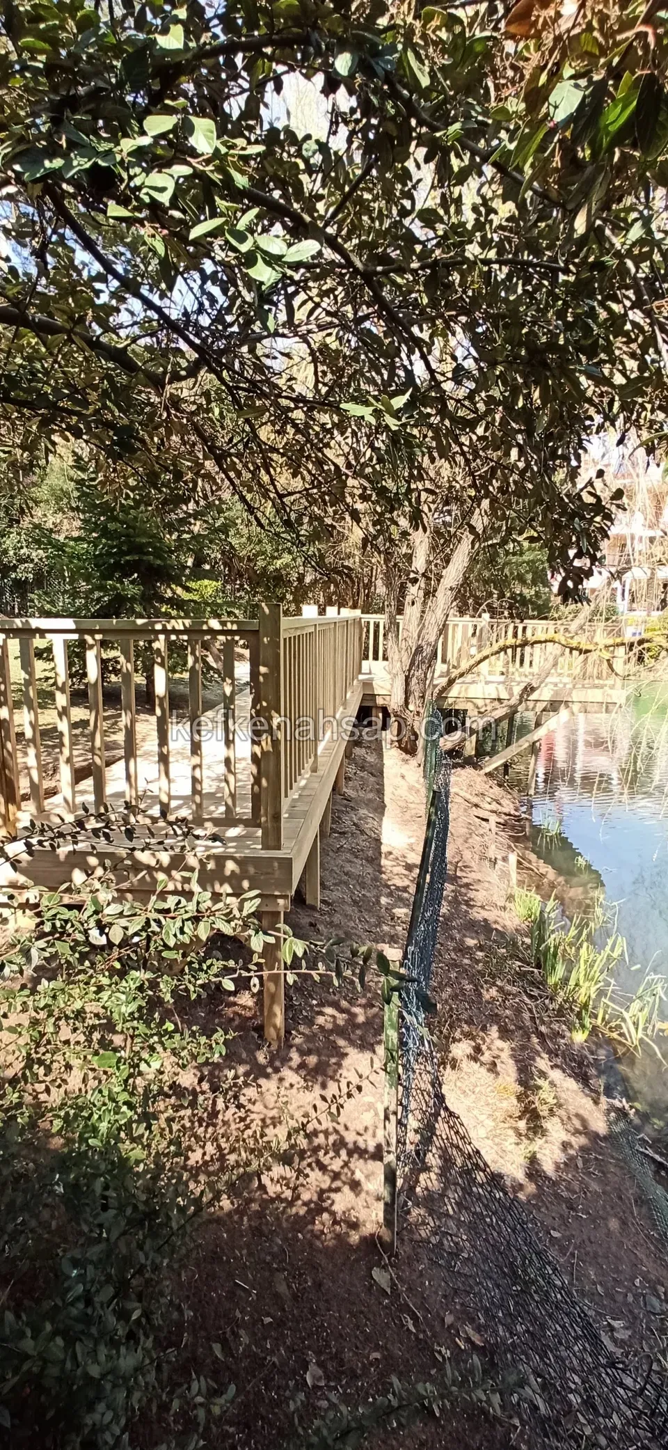 Светлое деревянное ограждение из горизонтальных реек вдоль кромки воды в тени деревьев