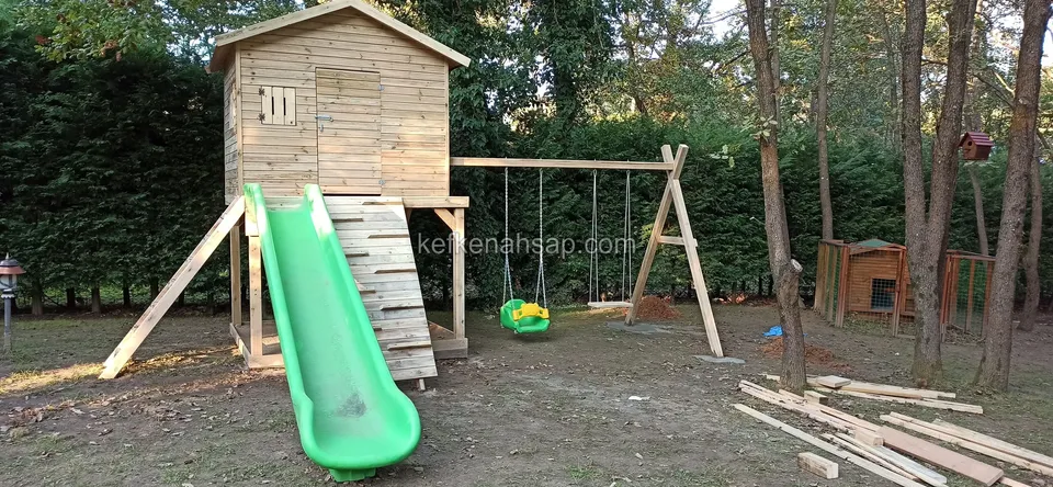 Ağaçların arasındaki bahçede, yüksekte duran ahşap oyun evi, yeşil kaydırak ve yan tarafta salıncak