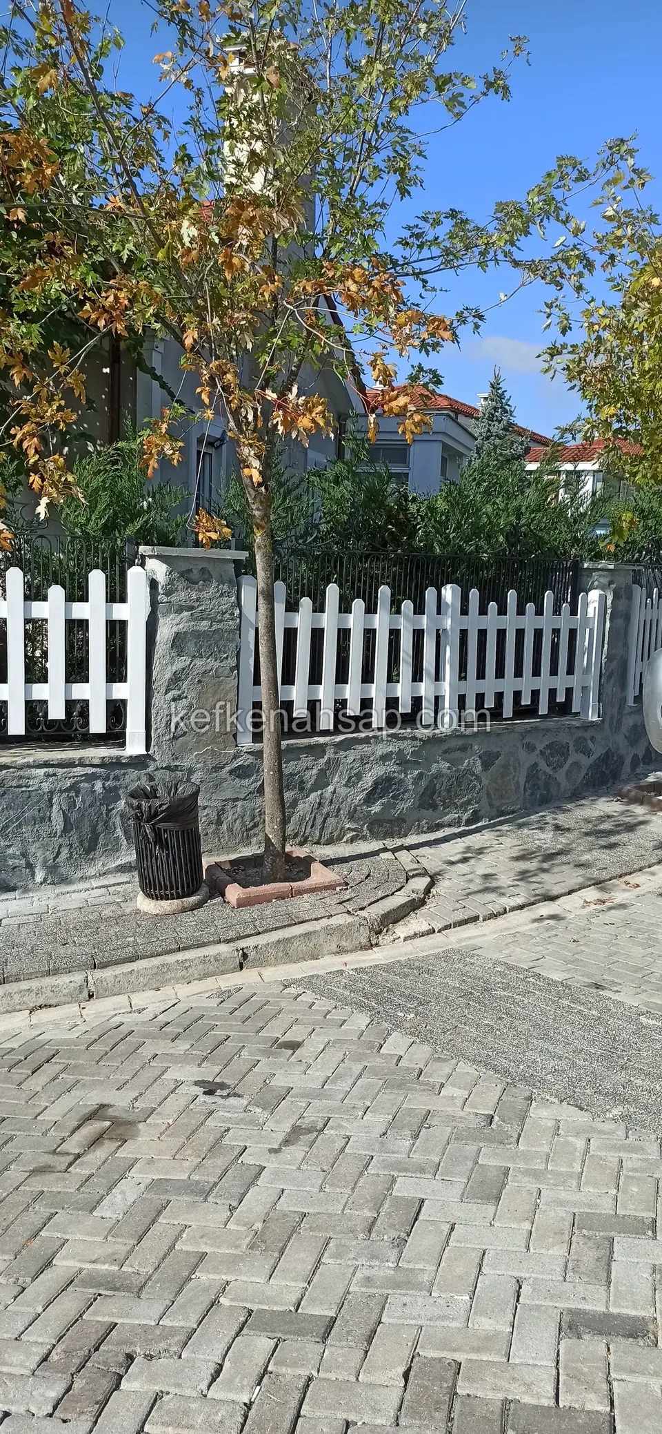 Sonbahar yapraklı ağacın dibinde, taş duvarın üstünde uzanan beyaz çıtalı çit ve çöp kovası