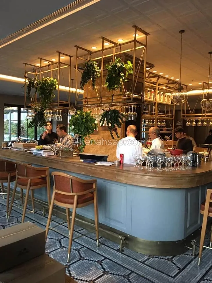 Masif ahşap tezgahlı kavisli bar bankosu ve önünde ahşap bar tabureleri bulunan restoran iç mekanı.