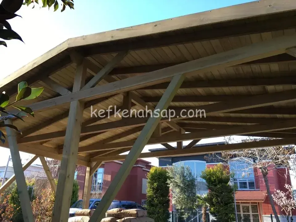 Alttan görünen ahşap çardak çatısı; kirişler, çapraz destekler ve tahta kaplama tavan, arkada kırmızı binalar ve ağaçlar