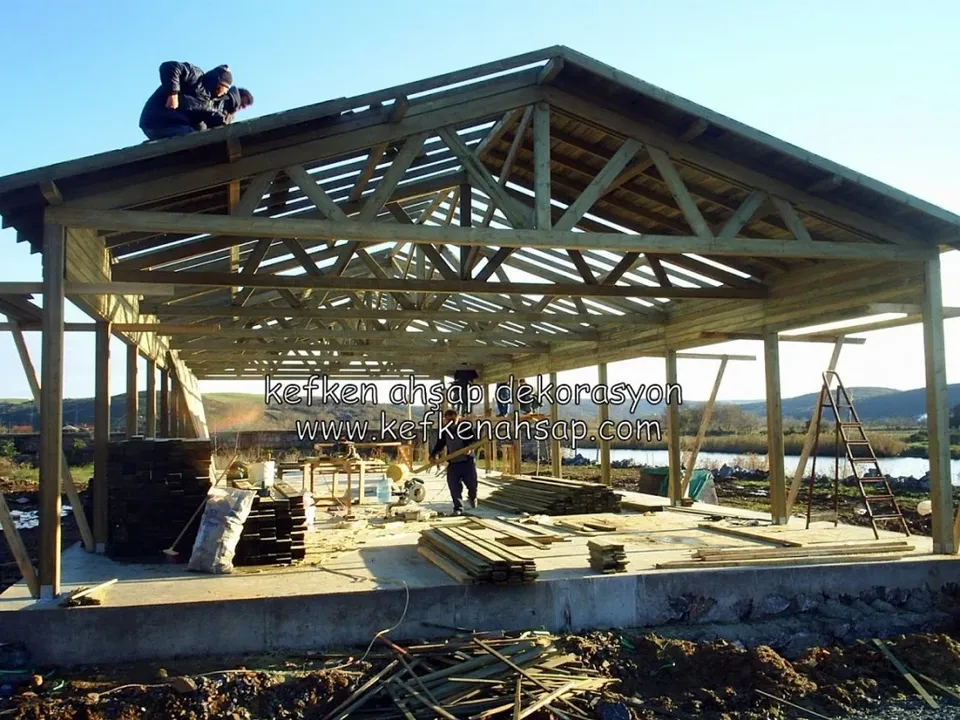 Beton temel üzerinde ahşap kolonlar ve çatı makasları; çatının üstünde iki işçi, zeminde istiflenmiş kereste var.