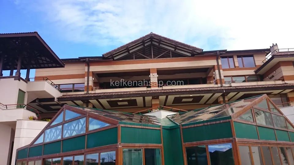 Wintergarten mit Holzrahmen, grünen Paneelen und Glasdach am Fuß eines mehrgeschossigen Gebäudes, darüber Holzüberstände