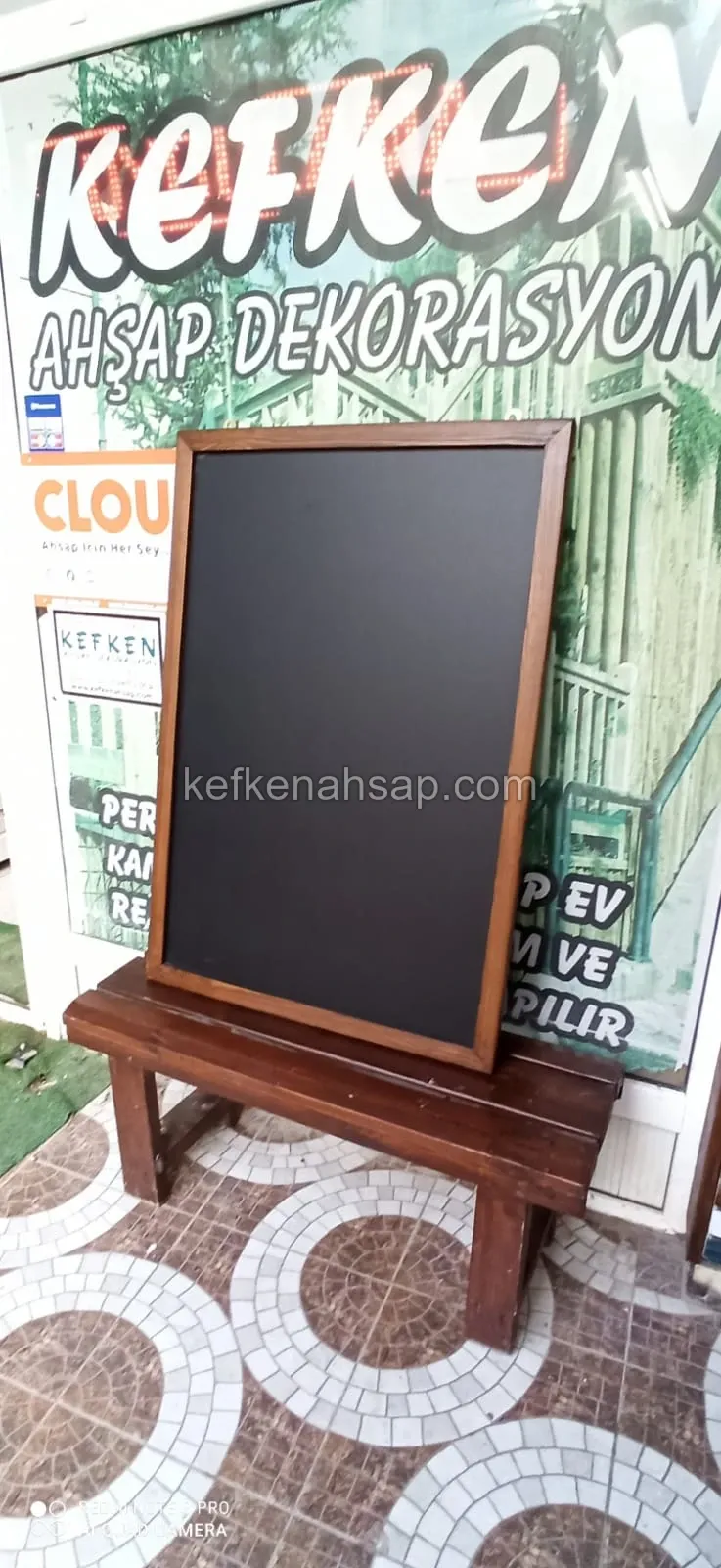 Black chalkboard in a dark brown wooden frame resting on a wooden bench at a shop entrance