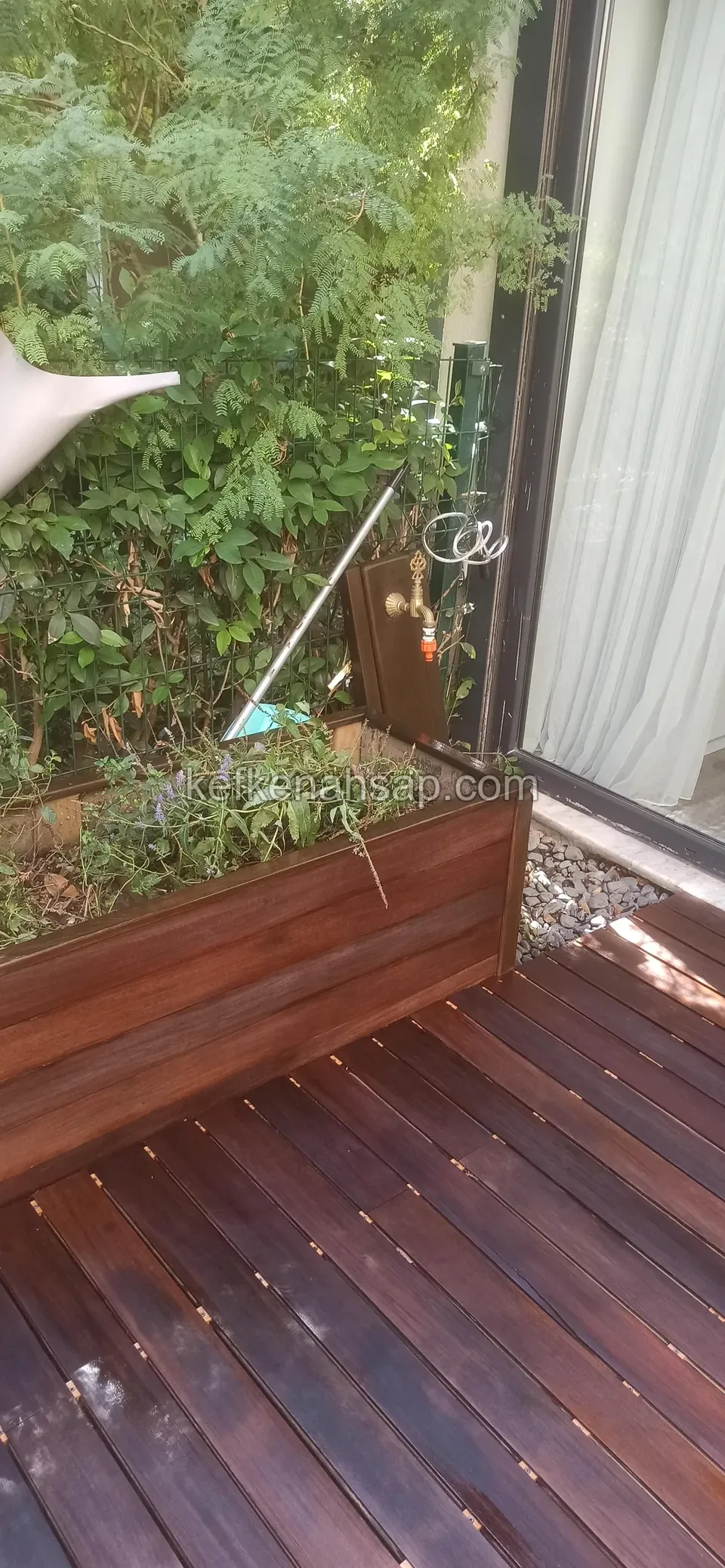 Dark red-brown deck beside a glass door in front of a timber planter box full of plants