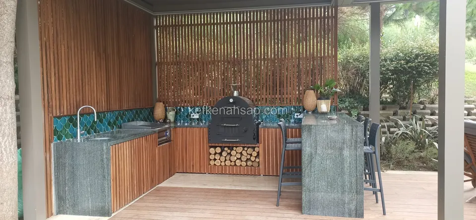 Dikey ahşap çıtalı duvar önünde ahşap kapaklı dolaplar, odun fırını ve bar tezgâhlı dış mutfak