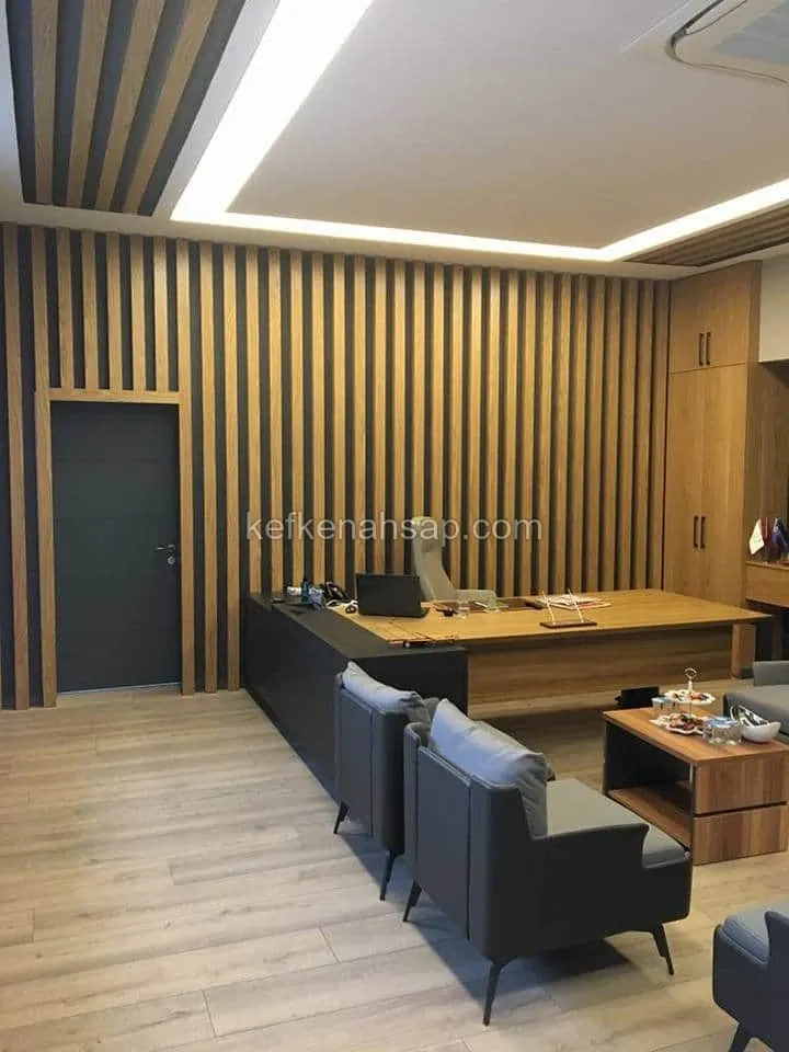 Dikey ahşap çıtalı duvar önünde L şekilli çalışma masası, gri koltuklar, sehpa ve gizli ışıklı tavan bulunan ofis