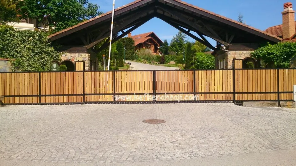 Çatılı taş ayaklı bir giriş yapısının önünde, metal çerçeveli dikey ahşap çıtalardan yapılmış geniş sürgülü kapı