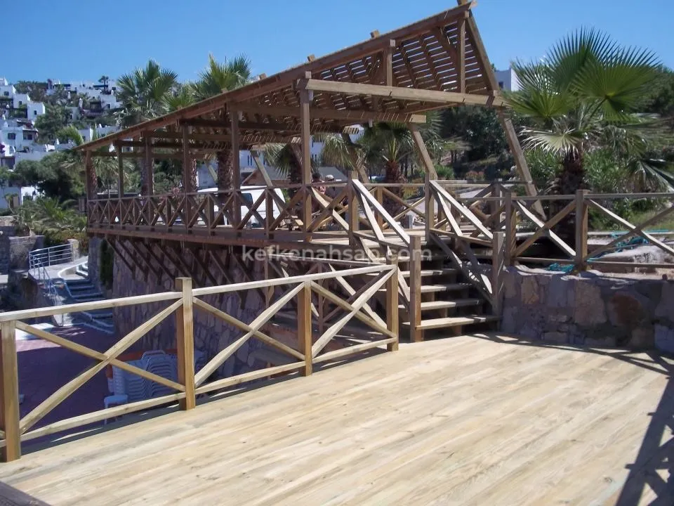 Taş duvar üstüne kurulu ahşap teras güvertesi, çapraz korkuluklar, merdiven ve çıtalı pergola çatısı.