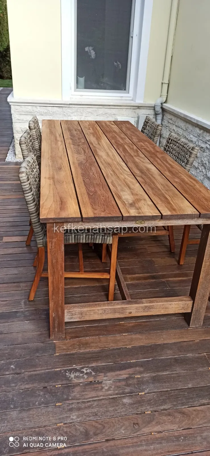 Long wooden dining table with wicker chairs on a wooden deck floor beside a stone-clad house