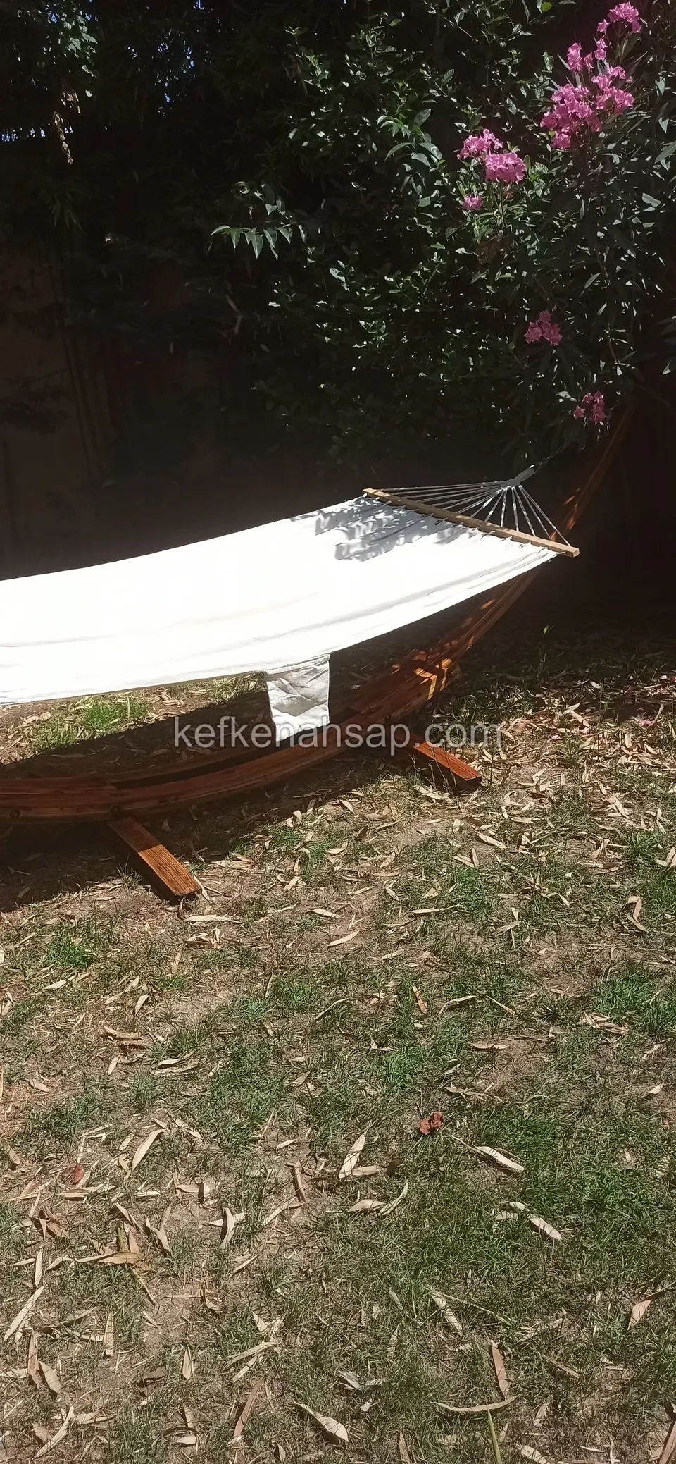 A cream-coloured hammock slung on a curved timber stand in a lawn garden, with flowering trees behind