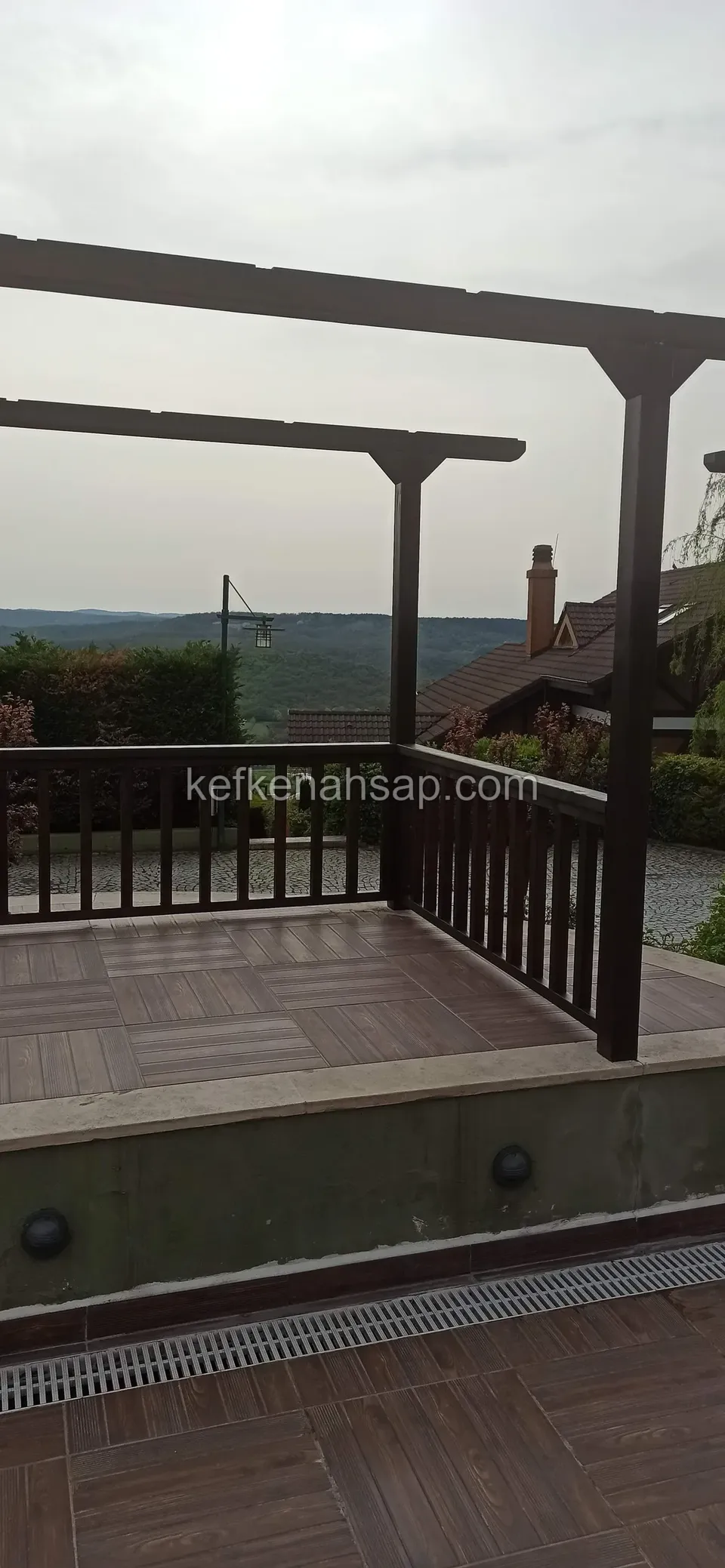 Ahşap korkuluklu platform, pergola direkleri ve kirişleri, ahşap görünümlü seramik zemin ve uzakta ağaçlık