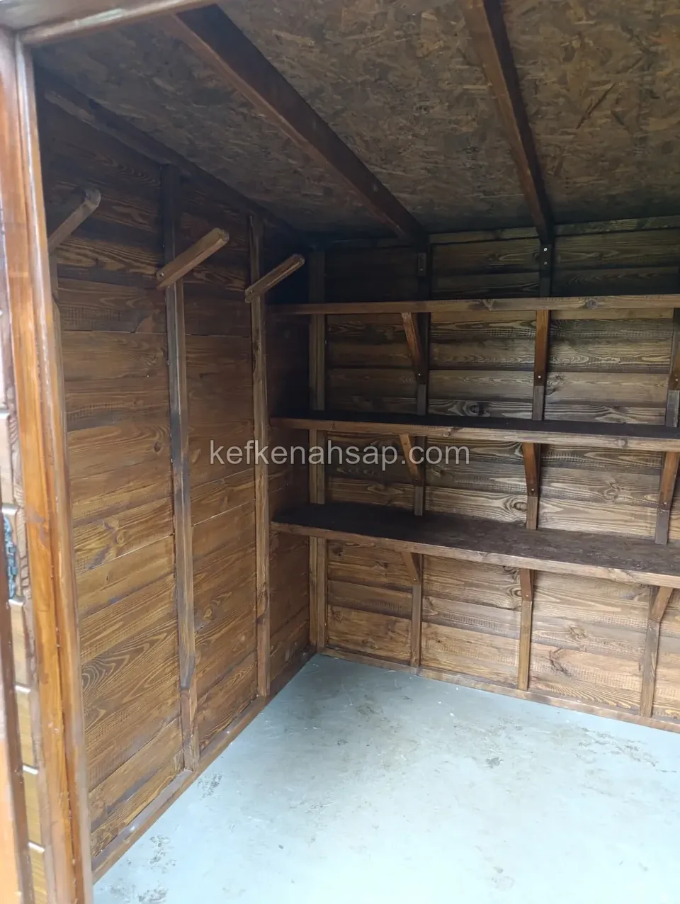 Innenraum einer Holzhütte mit braunem Lack, an der Seitenwand Kleiderstangen, an der Rückwand dreistufiges Regal, OSB-Decke