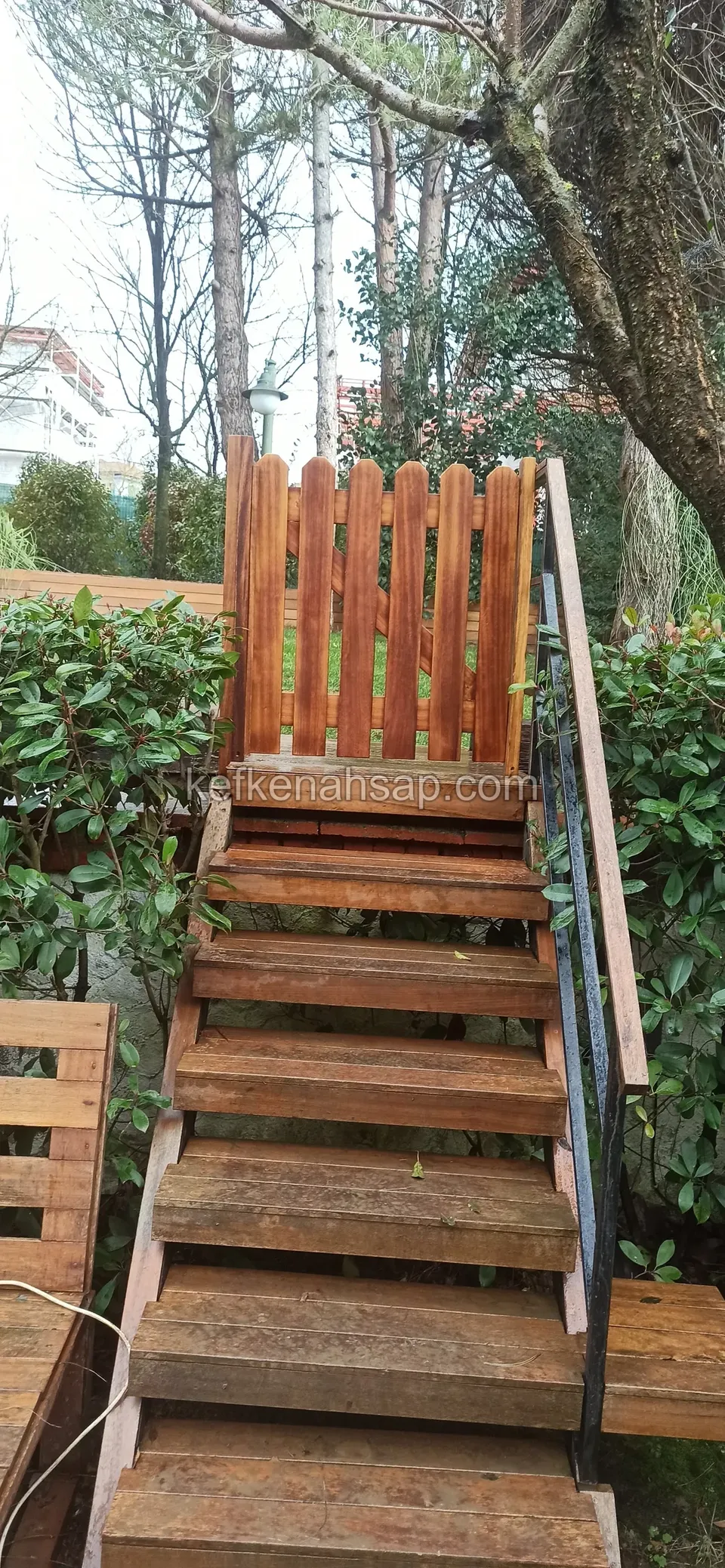 Yeşil çalılar arasında yükselen kahverengi tonlu ahşap basamaklar ve en üstte sivri uçlu ahşap bahçe kapısı