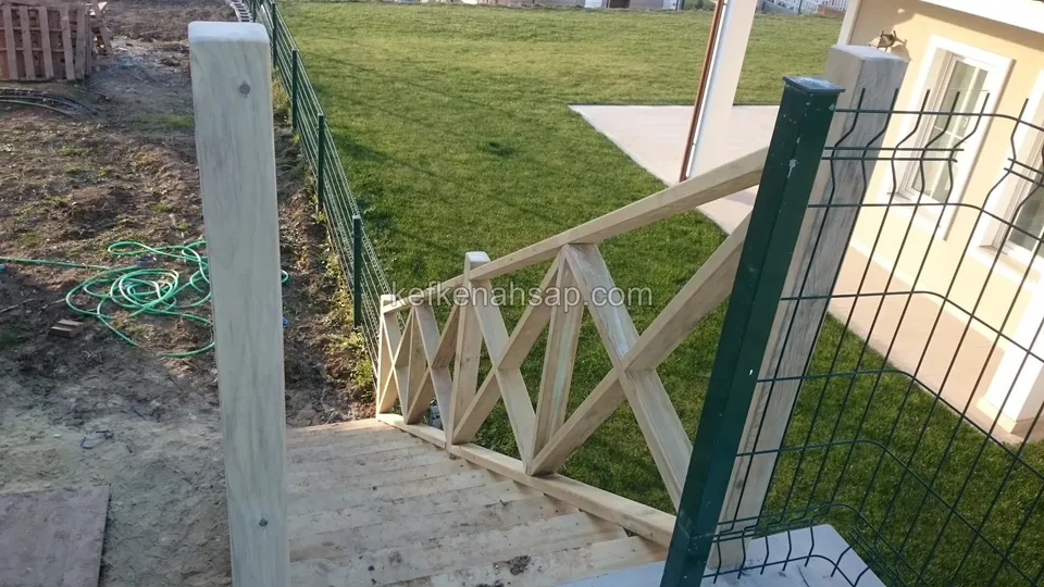 Çimliğe inen ahşap merdivenin kenarında X çaprazlı korkuluk, kalın bir direk ve yanında yeşil kaynaklı tel panel çit