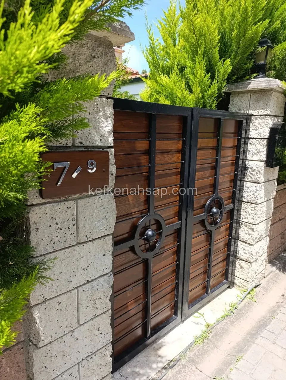 Taş sütunlar arasına monte edilmiş, siyah metal kasalı ve yatay ahşap panelli çift kanatlı bahçe kapısı.