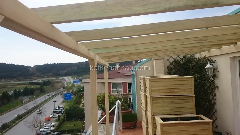 Terasta ahşap pergola iskeleti, önde üst üste dizilmiş ahşap saksılıklar, kafes panel ve aşağıda uzanan yol
