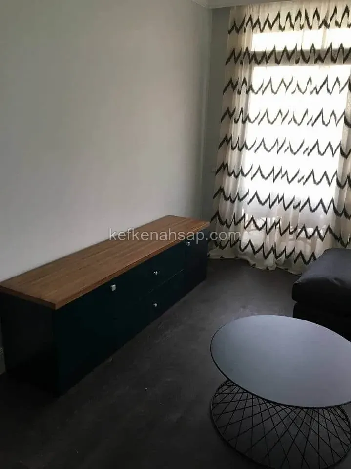 Oturma odasında koyu renk çekmeceli alçak dolap, üstünde bal rengi ahşap tabla; yanında yuvarlak sehpa ve desenli perde var