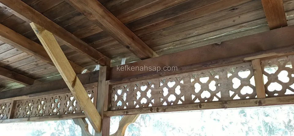 Kahverengi ahşap tavan kirişlerinin altında, geometrik motifli oyma kafes panel ve köşede eğik destek