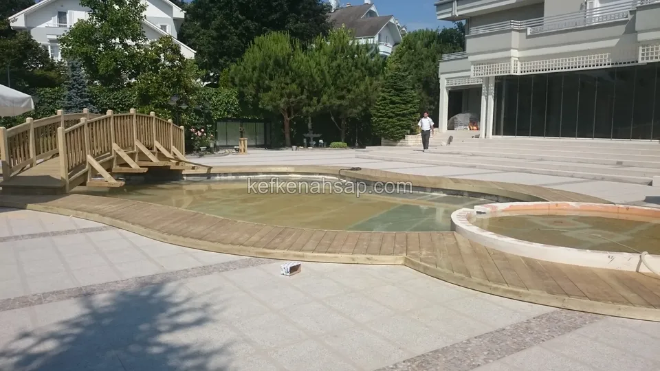 Alçak açıdan çekilmiş, deck kuşaklı sığ havuz ve uzaktaki ahşap köprü, arkada iki katlı bina
