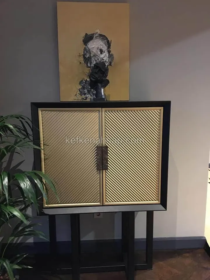 Schwarz gerahmter zweitüriger Schrank mit goldener Diagonalriffelung, darauf ein Bild mit gelbem Grund, daneben eine Pflanze