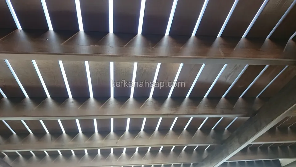 Alttan görünen koyu vernikli çıtalı pergola çatısı, ana kirişler ve aralardan süzülen gökyüzü