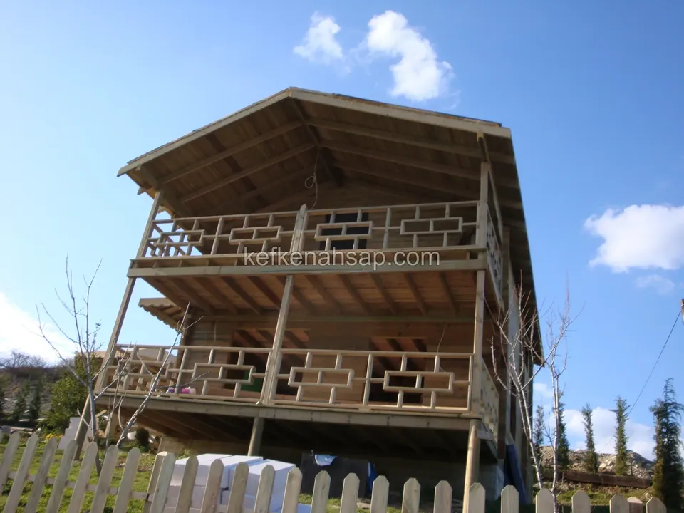 Çıtalı çitin arkasından, alttan bakılan iki katlı ahşap evin desenli balkon korkulukları