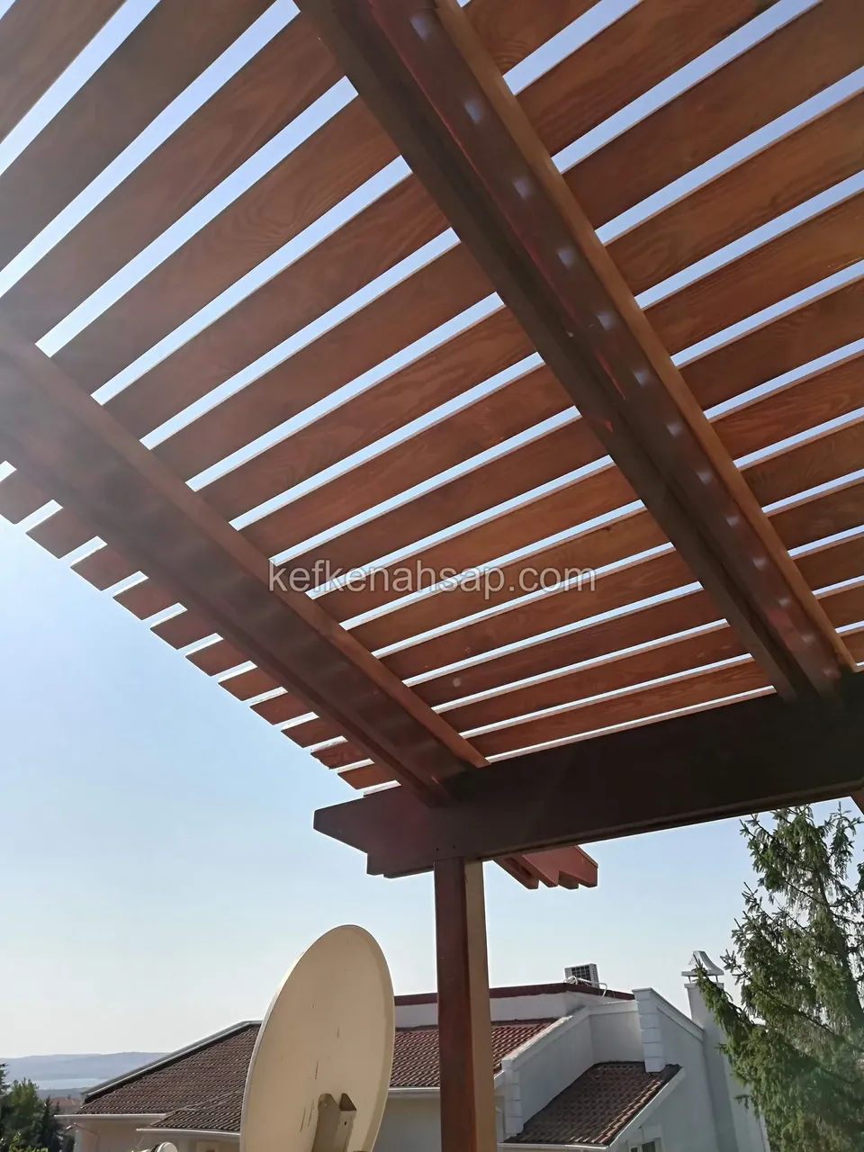 Alttan bakıldığında sık aralıklı vernikli ahşap pergola kirişleri, yanda uydu çanağı ve kıyıya bakan evler