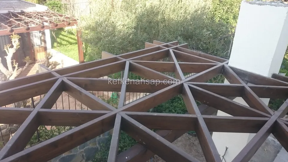 Alttan görünen üçgen kafesli vernikli pergola, çevrede bitkiler, tuğla ve beyaz sıvalı kolonlar, yanda başka bir pergola