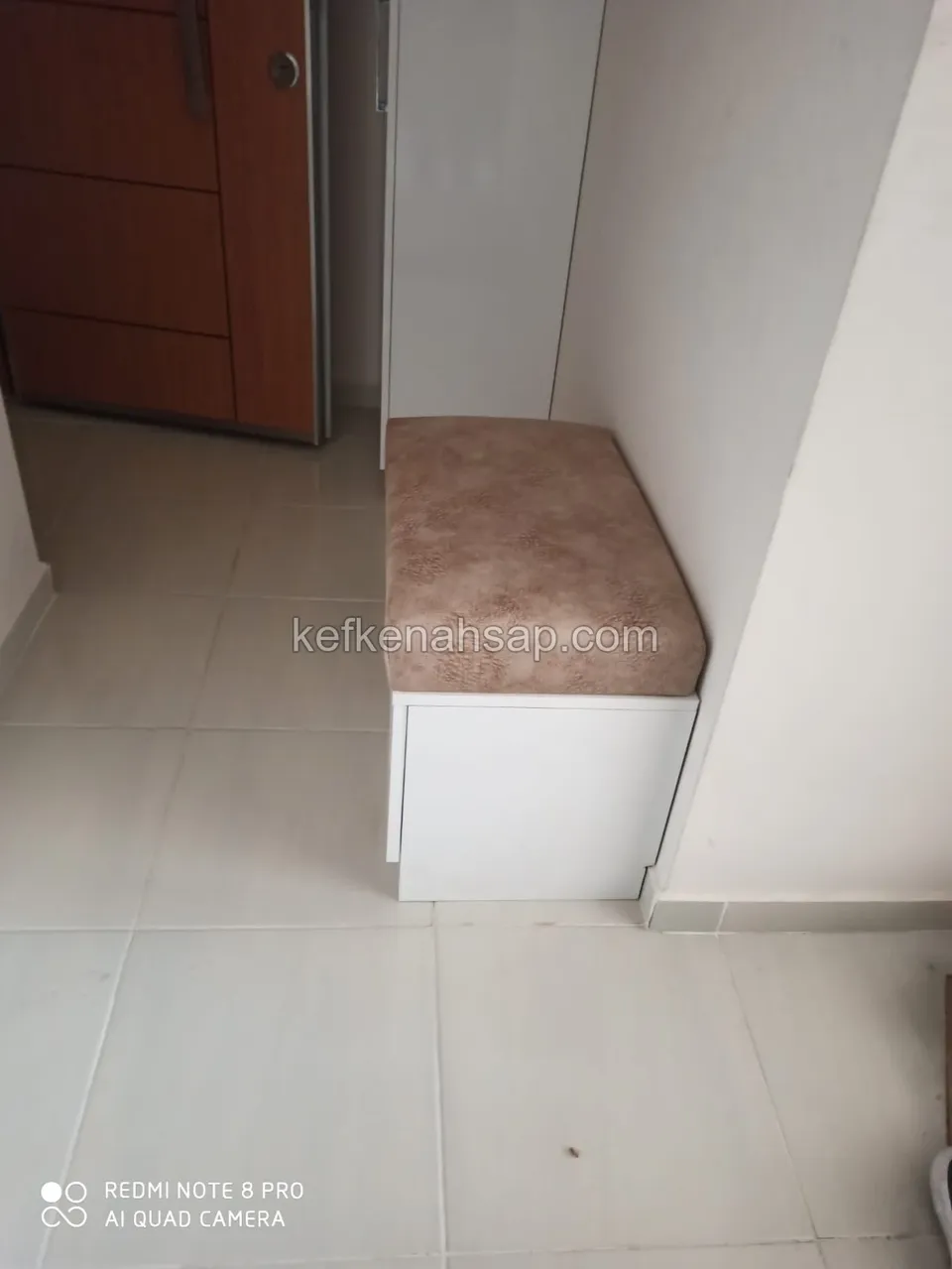 White storage bench with a beige upholstered seat in an entryway beside a white cabinet, wood-look door behind