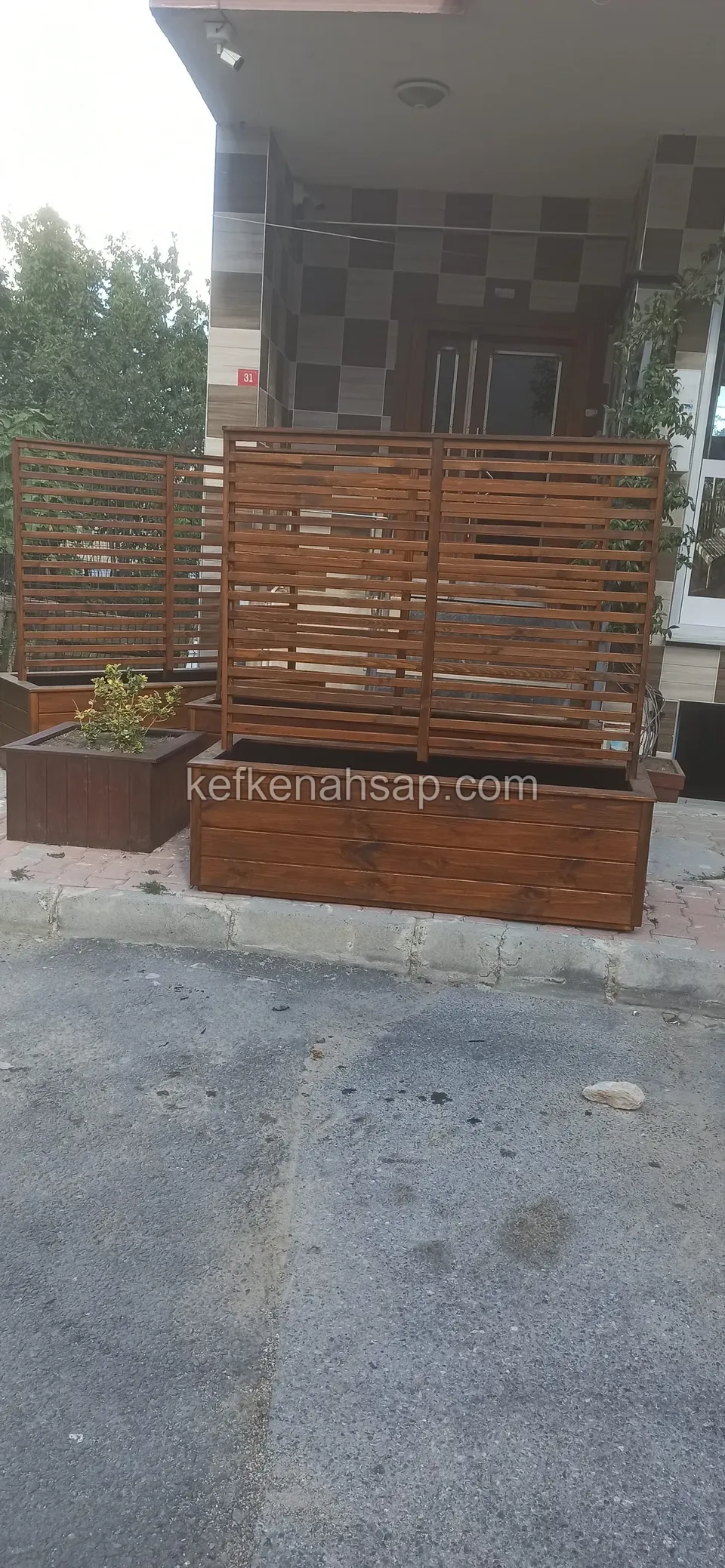 Asfalt yolun kenarında bina girişinin önünde duran, yatay çıtalı paravanlı ahşap çiçeklikler; arkada ekose karolu giriş