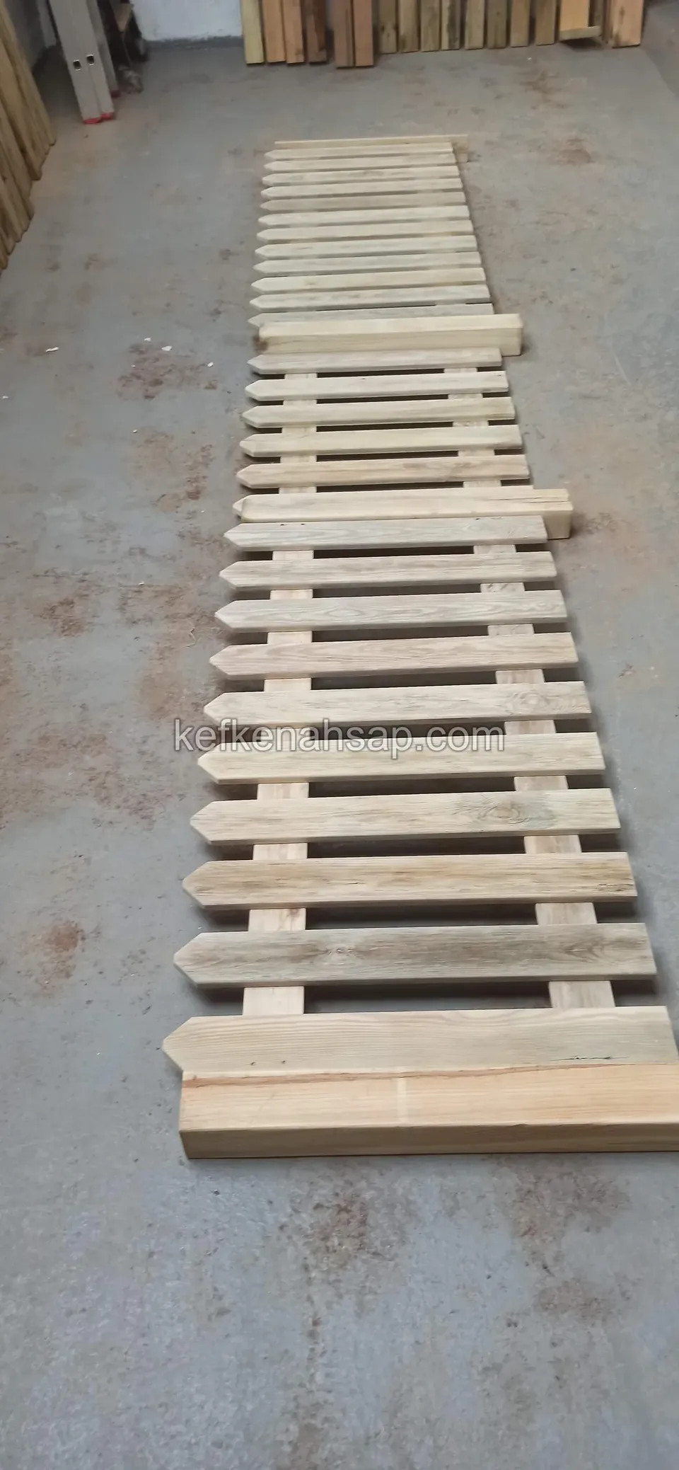 A light pointed-slat fence panel laid lengthwise on the grey floor of a workshop