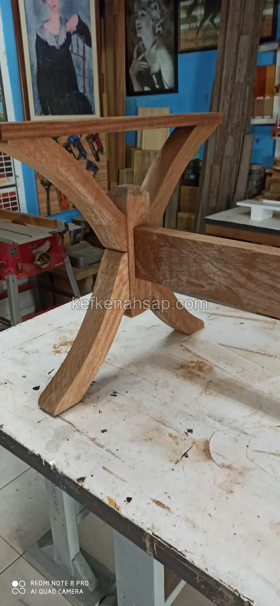 Auf einer weißen Werkbank liegt ein Holztischfuß, dessen geschwungene Arme sich zu einem X treffen, dazu ein langer Balken