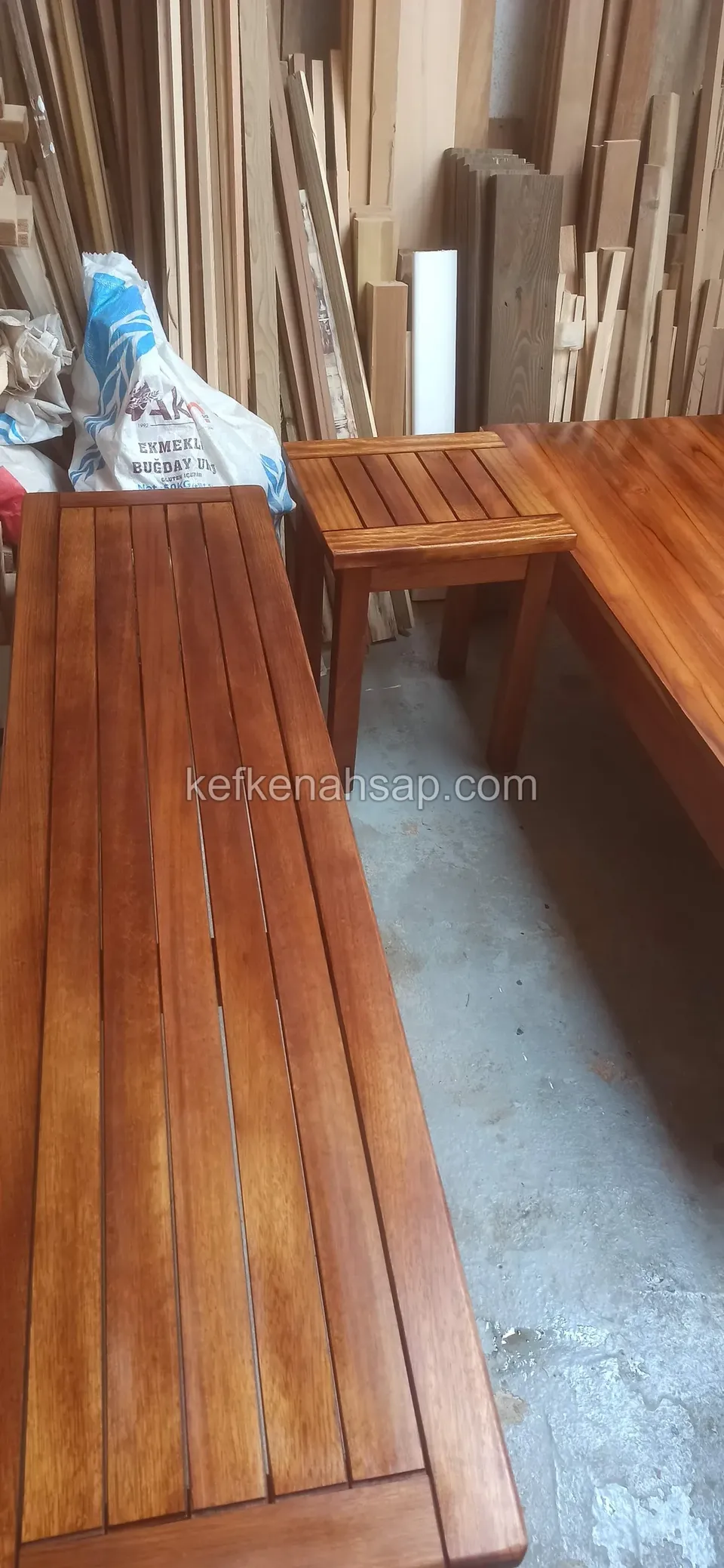 On a workshop floor, a long slatted timber bench and a small slatted table beside it in reddish varnish