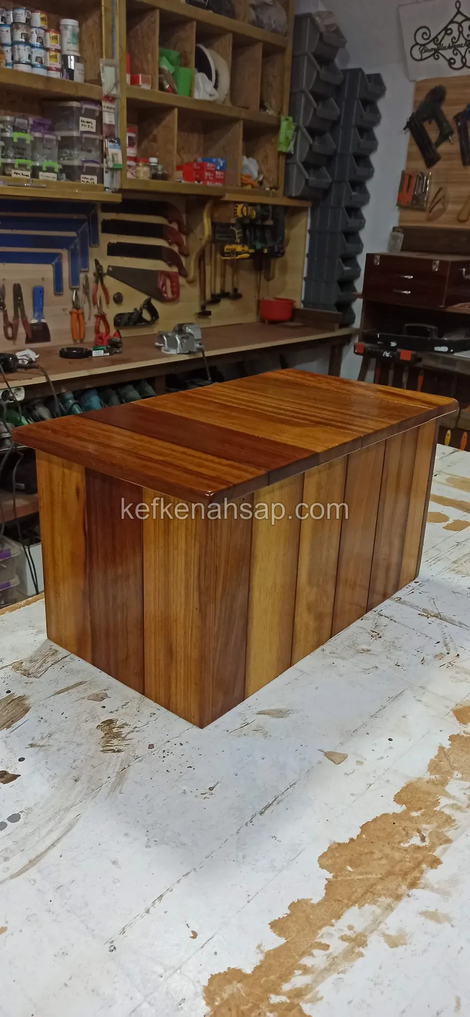 In a workshop in front of a tool board, a box-shaped timber chest with vertical strip sides and a glossy top