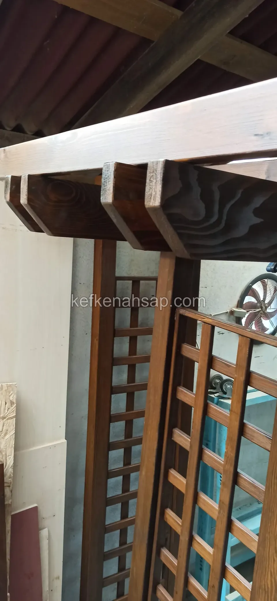 Atölyede kare gözlü ahşap kafes panelin yanında, üstte desenli kesilmiş kiriş uçları ve koyu cilalı direk