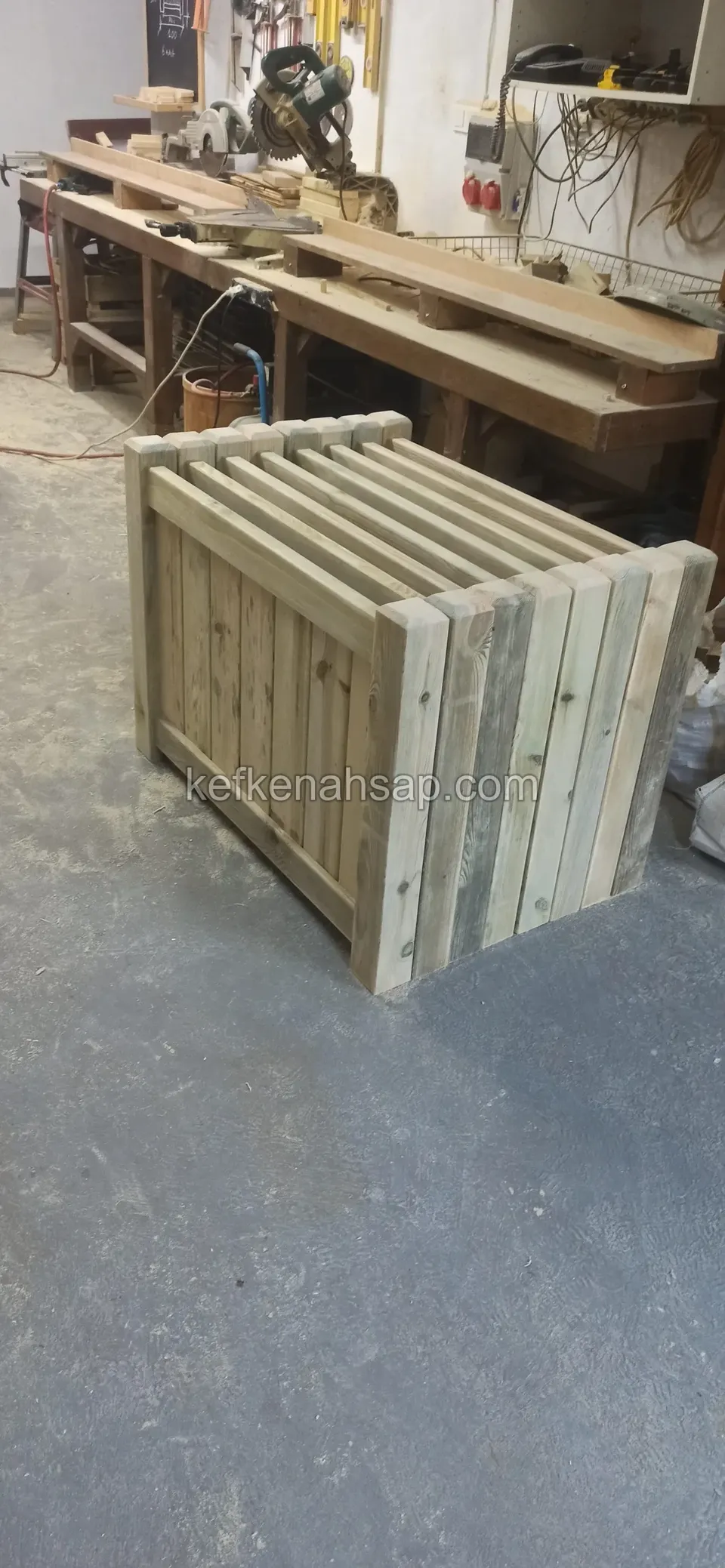 Square timber box with slatted sides being built in the workshop, with a work bench behind
