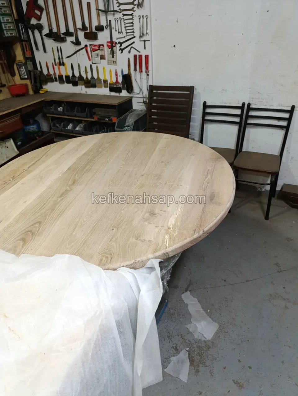 Helle runde Tischplatte in der Werkstatt, teilweise mit weißem Tuch bedeckt, an der Wand hängendes Werkzeug und Stühle