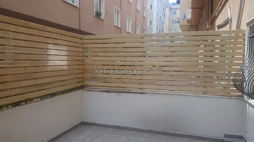 Beyaz sıvalı avlu duvarının üstünde L biçiminde uzanan açık tonlu yatay çıtalı ahşap paravan, altta karo zemin