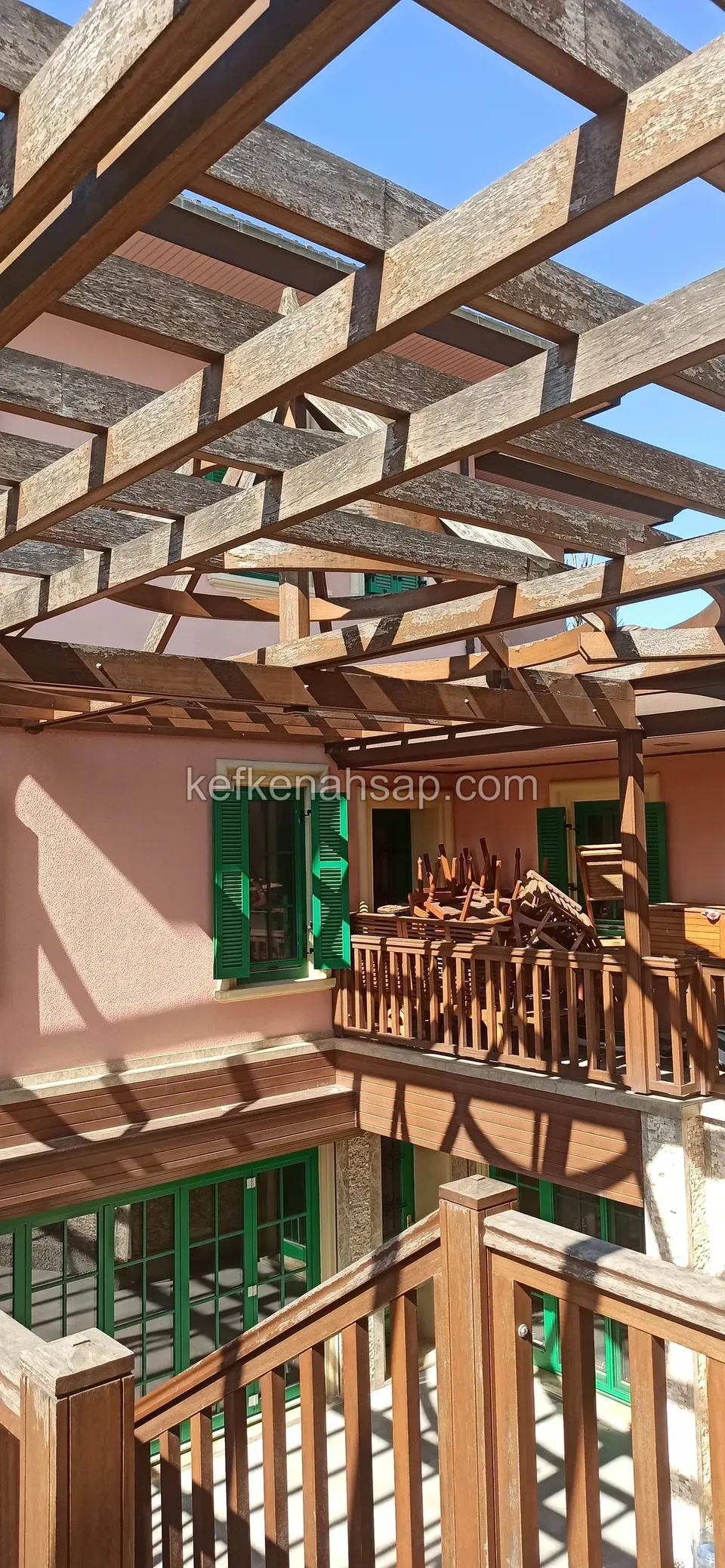 Yeşil panjurlu pembe binanın avlusunda, ahşap balkon korkulukları üzerinde uzanan kahverengi pergola