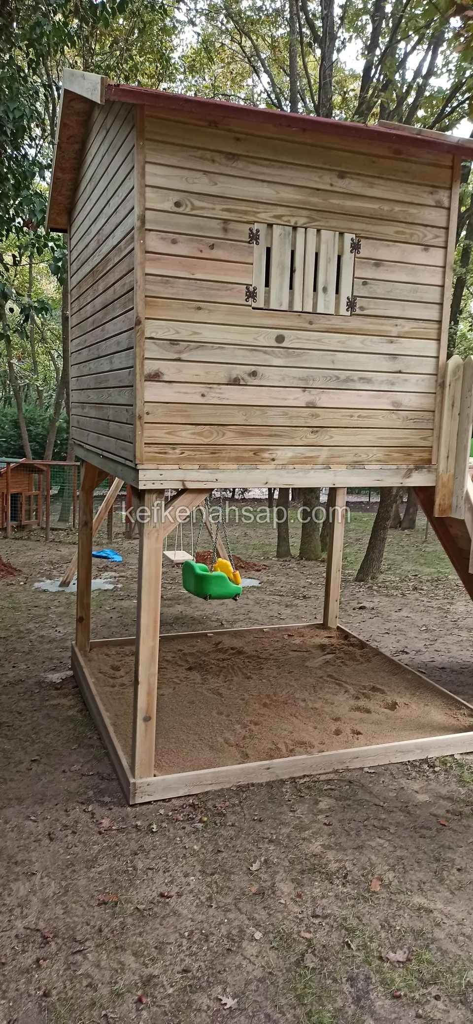 Ayaklar üzerinde yükseltilmiş ahşap oyun evi; altında çerçeveli kum havuzu ve asılı bir salıncak