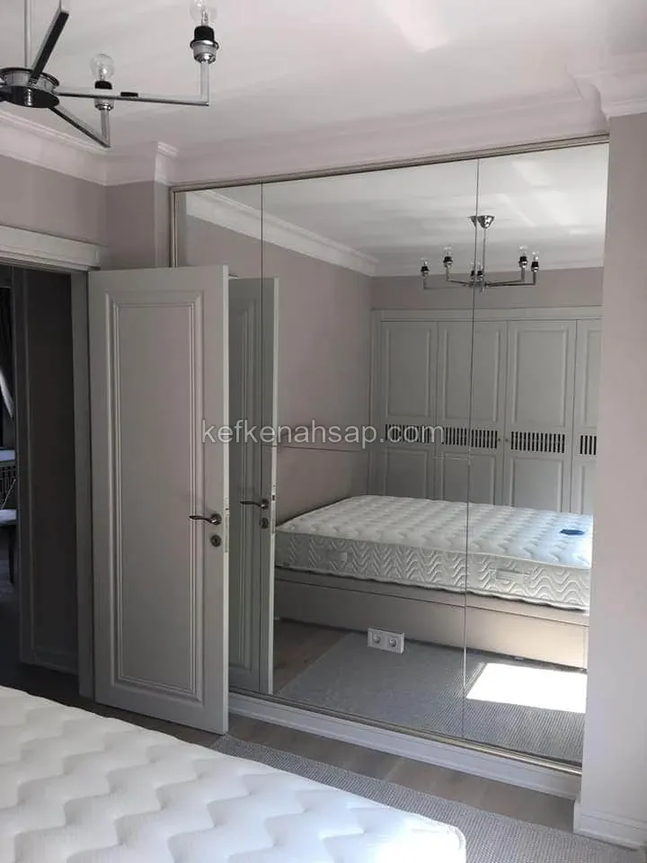 Yatak odasında boydan boya ayna kaplı dolap yüzeyi, açık gri boyalı çerçeveli kapı ve aynada görünen yatak