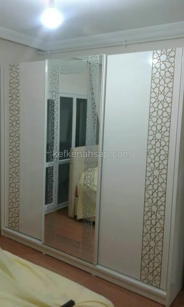 Yatak odasında üç kapaklı beyaz sürgülü gardırop; ortada ayna, yanlardaki kapaklarda altın renkli geometrik desen şeritleri